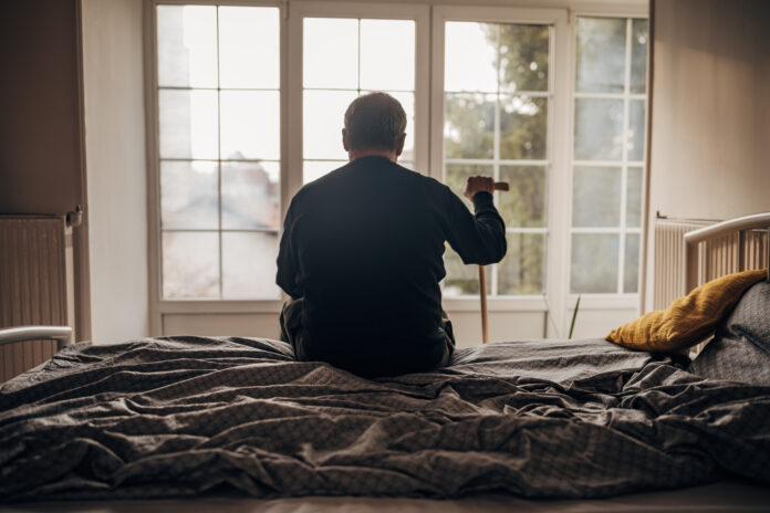 An elderly man is sitting on the bed with his back to the camera