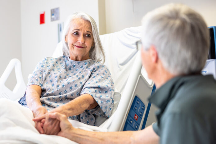 A senior woman in the hospital room recovering, while her husband supports her.